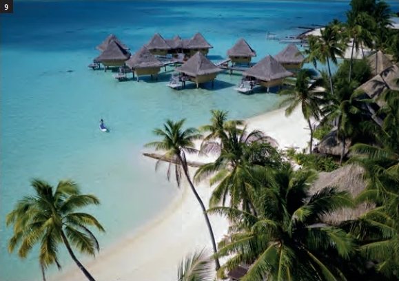 Luftaufnahme der Overwater-Bungalows in der türkisfarbenen Lagune und des von Kokospalmen gesäumten Strandes.
