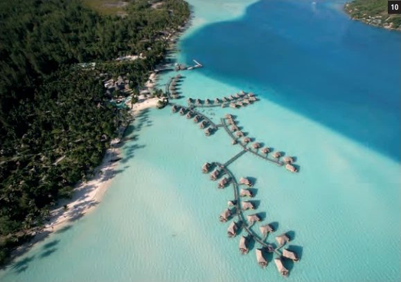 Luftaufnahme des Bora Bora by Pearl Resorts mit seinen Overwater-Bungalows in der türkisfarbenen Lagune und dem bewaldeten Motu.