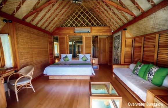 Innenansicht eines hölzernen Overwater-Bungalows mit Doppelbett, Sofa mit grünen Kissen und Couchtisch mit Glasplatte.