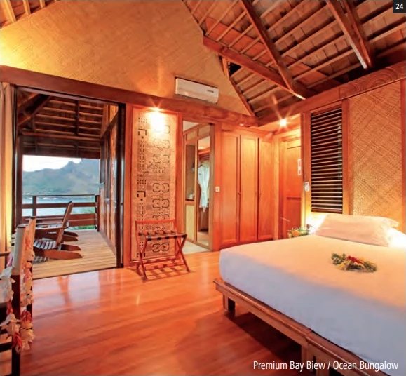 Holzverkleideter Innenraum eines Bungalows mit grossem Bett und offener Terrasse mit Blick auf Bucht und Berge.