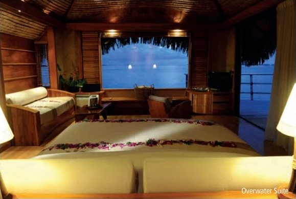 Innenansicht einer Overwater-Suite in der Abenddämmerung: mit Blumen dekoriertes Bett vor einer zur Lagune hin geöffneten Glasfront.