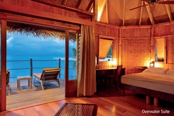 In Holz gehaltenes Interieur einer Overwater Suite mit Bett, Schreibtisch und zur Lagune hin offener Terrasse.