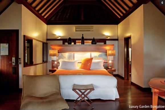 Zimmer eines Luxury Garden Bungalows mit grossem weissem Bett, orangefarbenem Kissen und dunkler Holzdecke.