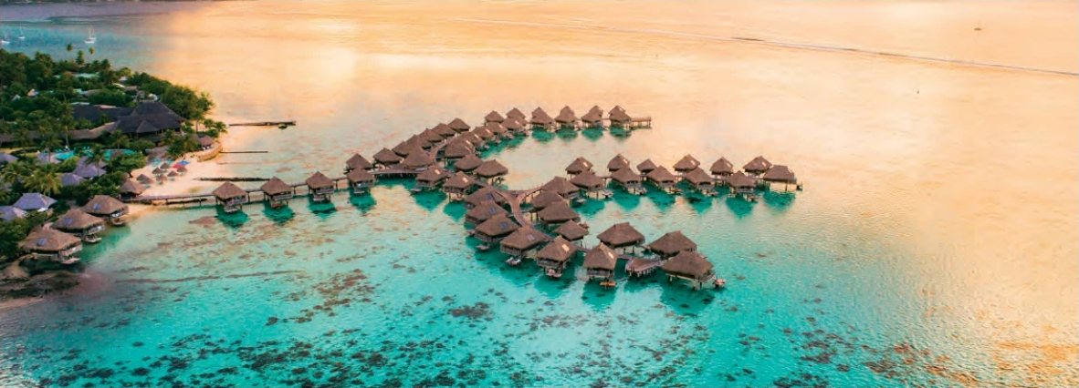 Luftaufnahme eines Hotels mit Überwasser-Bungalows in einer türkisfarbenen Lagune bei Sonnenuntergang.