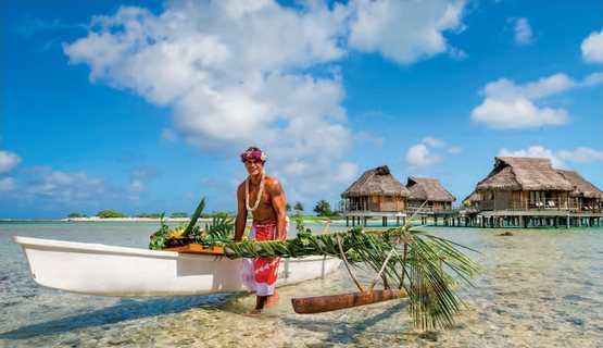 Ein Polynesier mit Blumenkranz schiebt ein mit Palmwedeln geschmücktes Auslegerkanu durch die Lagune, dahinter Overwater-Bungalows.