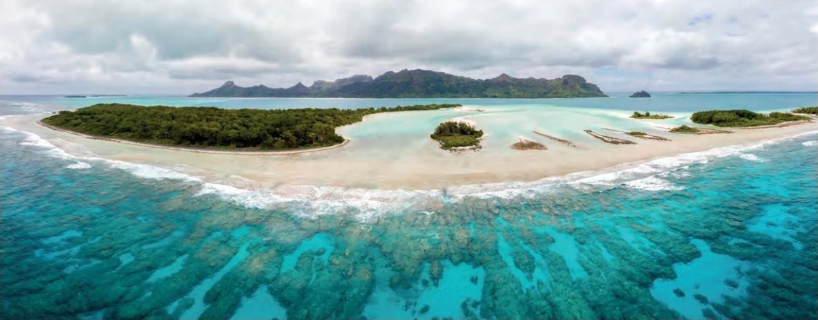 Aerial view of a motu fringed with white sand and a turquoise coral reef, with a mountainous island on the horizon.