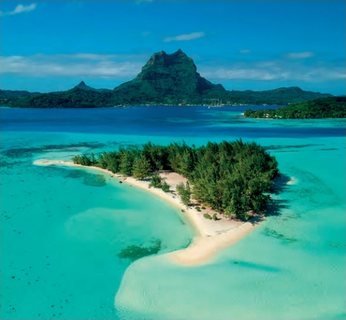 Luftaufnahme eines bewaldeten Motu mit weissem Sandsaum in einer türkisfarbenen Lagune, im Hintergrund die Bergsilhouette von Bora Bora.