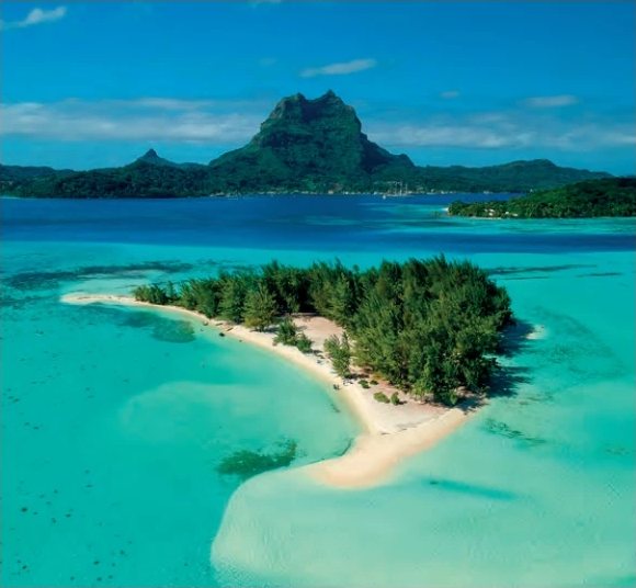 Aerial view of a wooded motu fringed with white sand in a turquoise lagoon, with the mountainous silhouette of Bora Bora in the background.