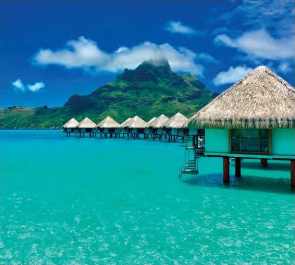 Thatched overwater bungalows on a turquoise lagoon, with a lush green mountain in the background.